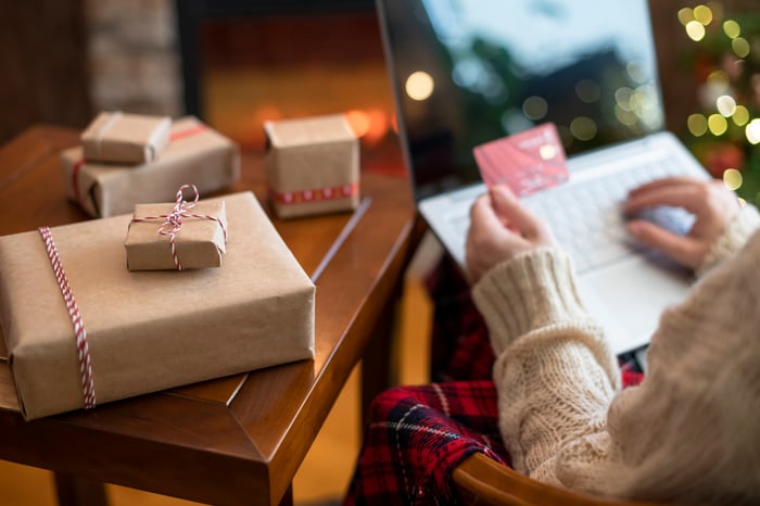 Christmas.-Woman-in-sweater-holding-credit-card-using-laptop-for-making-order-sitting-at-table-with-packaging-gift-near-fireplace-and-christmas-tree.-Online-shopping-concept-1286677721_2125x1416